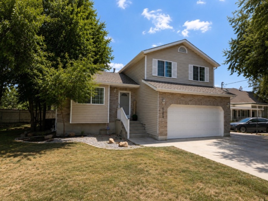 Tri-level home with tan siding, brick accents, an attached two-car garage, and mature trees over a green front lawn.